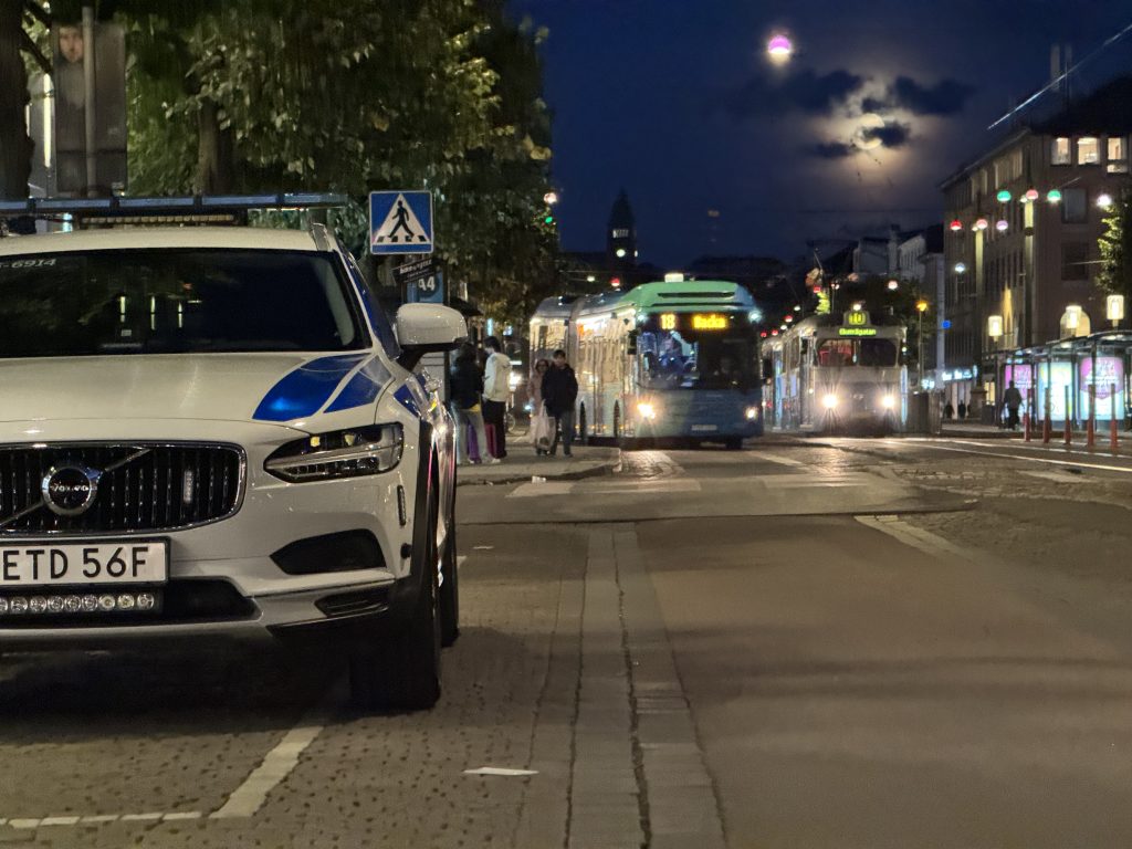 En vit bil nattetid som står parkerad med en buss och spårvagn i bakgrunden