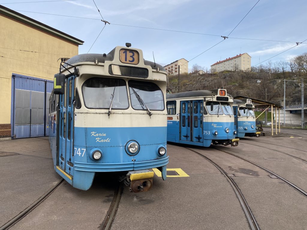tre blåvita spårvagnar utanför en gul depåbyggnad.