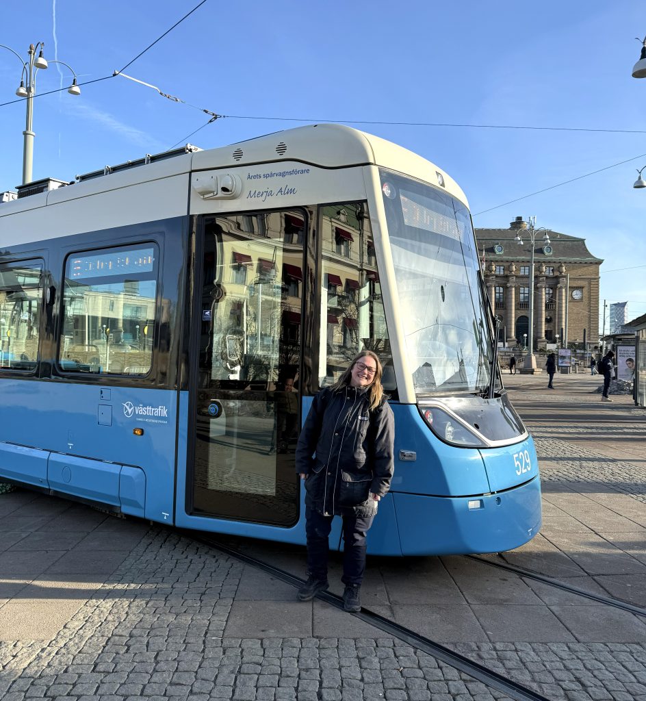 En glad spårvagnsförare framför en blåvit spårvagn.